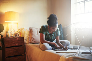 Eine junge Frau lernt für ihr Fernstudium auf ihrem Bett im Schneidersitz.