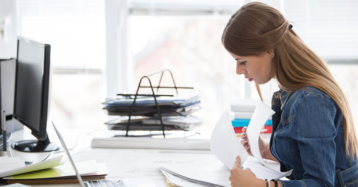 Junge Frau sitzt am Rechner und blättert verschiedene Unterlagen durch.