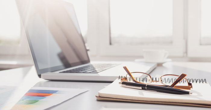 Neben einem aufgeklappten Laptop liegt ein Notizbuch mit einer Brille oben drauf.