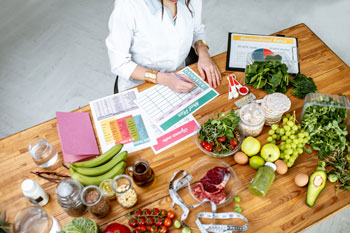 Eine Ernährungsberaterin sitzt am Schreibtisch mit vielen Unterlagen zu Auswertung, umgeben von buntem Obst und Gemüse