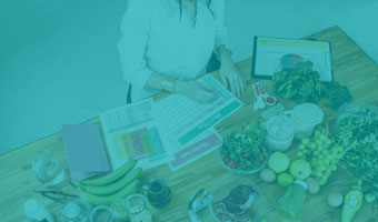 Eine Ernährungsberaterin sitzt am Schreibtisch mit vielen Unterlagen zu Auswertung, umgeben von buntem Obst und Gemüse