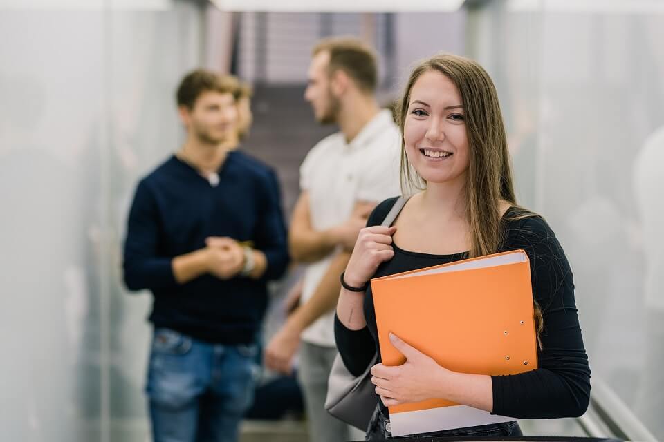 Masterstudierende der Sozialen Arbeiten stehen im Flur waehrend Studentin mit ihren Unterlagen in die Kamera laechelt